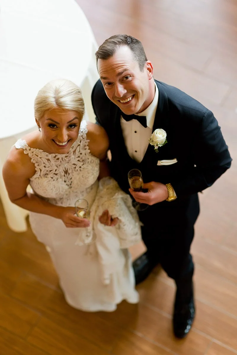 A joyful couple in wedding attire, viewed from above. The bride wears a lace gown, the groom a black tuxedo. Both holding champagne flutes, smiling warmly.