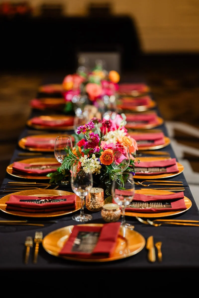 A lavishly set table with vibrant floral centerpieces, gold plates, and red napkins, creating an elegant and festive atmosphere. Wine glasses sparkle under soft lighting.