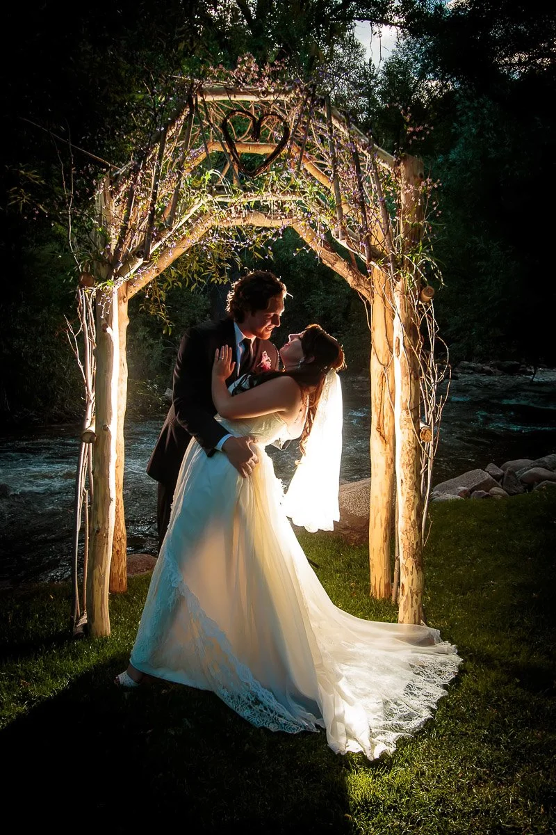 A bride and groom share a tender embrace under a softly lit, wooden arch adorned with branches. The scene is romantic and serene, with a gentle Boulder Creek in the background.