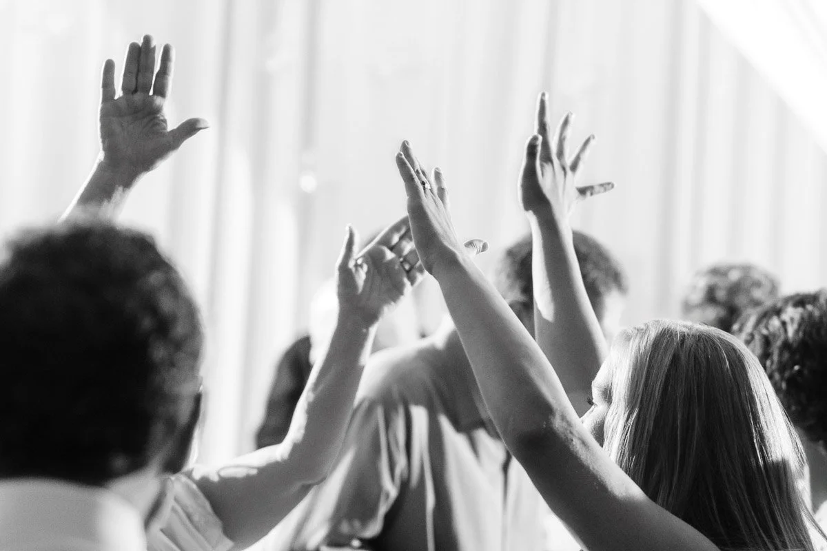 Black and white image of people with hands raised, as if dancing or celebrating, set against soft drapery. The mood is lively and joyful.