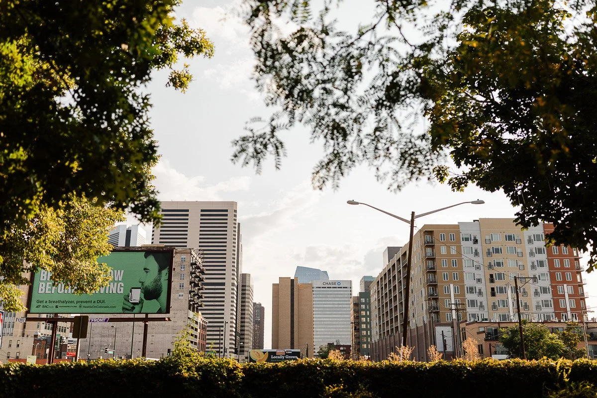 Cityscape with modern buildings framed by leafy trees. A prominent billboard with green hues is on the left. The scene exudes an urban, lively vibe.