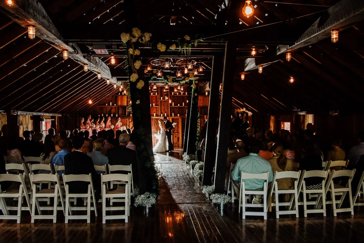 Bride and groom stand hand-in-hand on green grass, with a rustic wooden wall and animal skulls behind. The bride in an elegant floral gown holds white flowers, while the groom wears a classic black suit, both exuding a serene and timeless elegance.