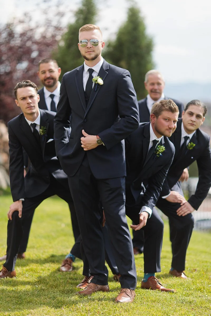A group of six men in dark suits pose playfully on grass, with one man in sunglasses standing confidently in the center. The mood is joyful and celebratory.