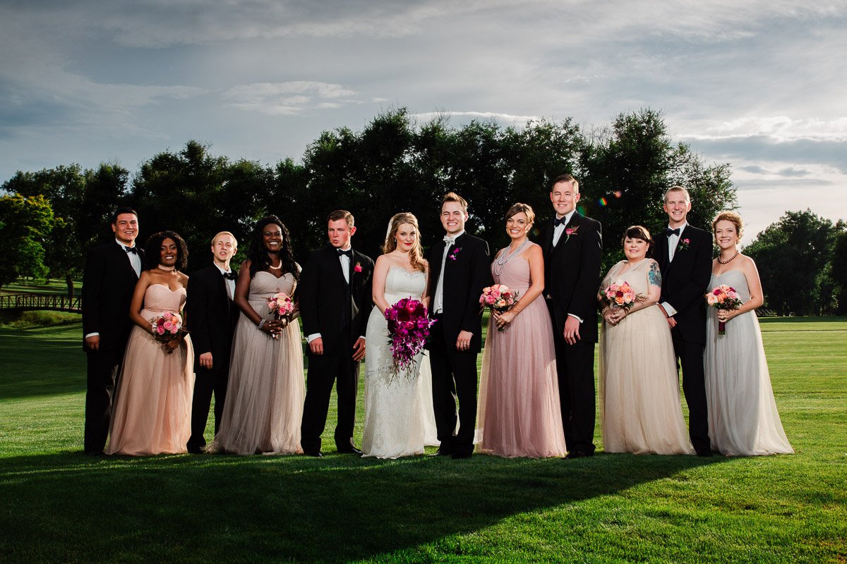A wedding party stands on a lush lawn with a backdrop of trees. The bride in white and bridesmaids in pastel dresses hold bouquets, exuding joy and elegance.