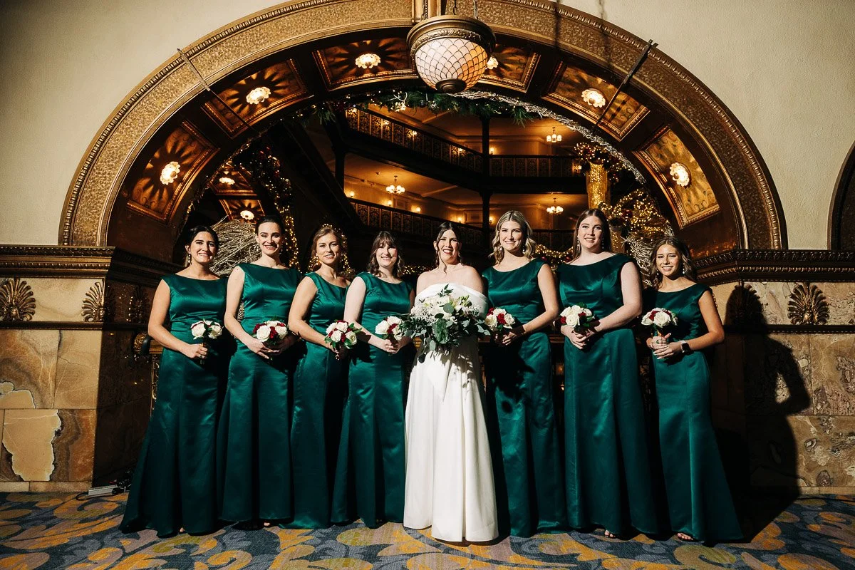 A bride in a white gown stands with seven bridesmaids in teal dresses, holding bouquets. They're under an ornate arch at the Brown Palice hotel, with warm lighting, conveying elegance.