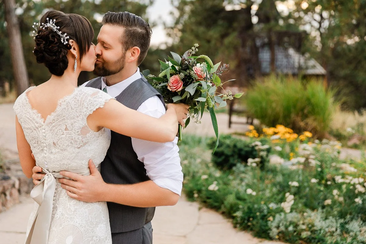 A bride and groom share a kiss outdoors. The bride wears a lace dress and holds a bouquet of pink flowers. The scene is surrounded by greenery and flowers, conveying romance and joy.