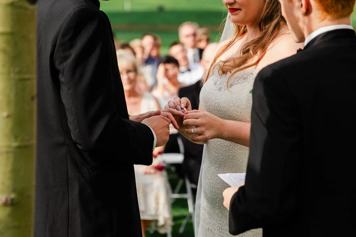 Bride and groom exchange rings during an outdoor wedding ceremony. Guests watch in the blurred background, conveying a joyful and intimate atmosphere.