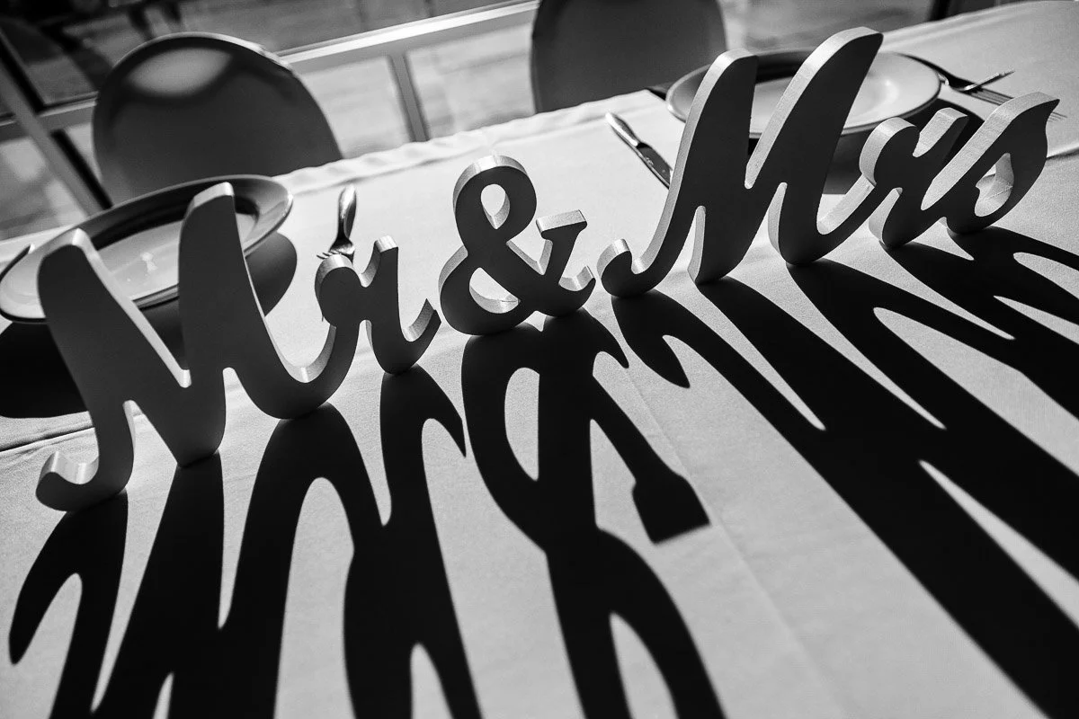Elegant "Mr & Mrs" sign casts bold shadows on a white tablecloth, flanked by empty plates and cutlery in soft sunlight, creating a romantic ambiance.