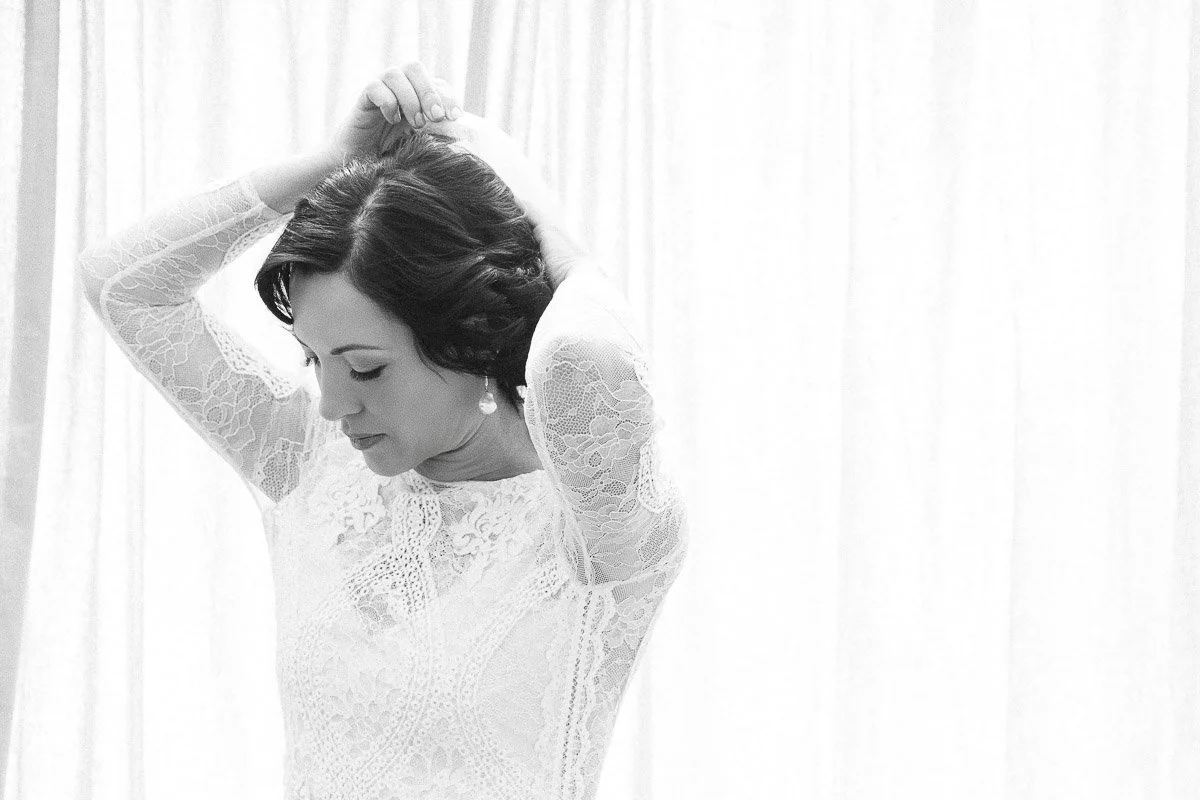 Black and white image of a woman in a lace dress, arms raised, adjusting her hair against a softly draped curtain. The mood is serene and contemplative.