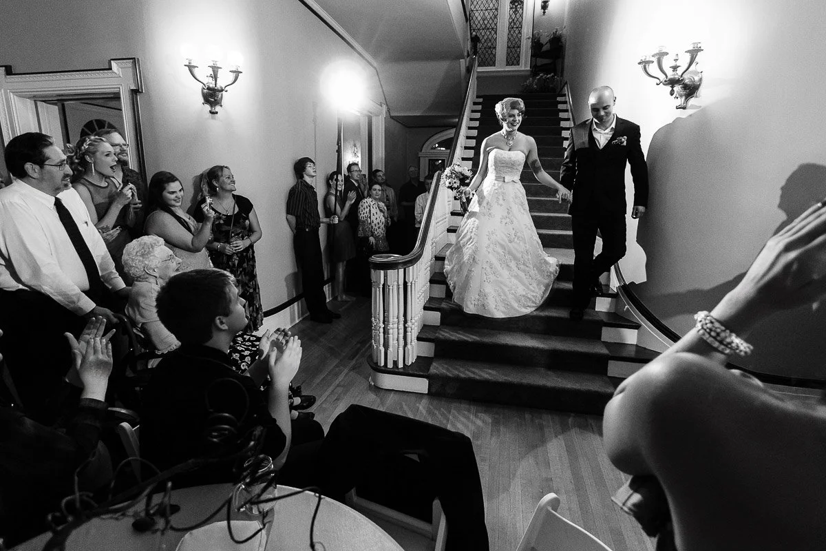 A bride in a white gown and groom in a dark suit descend a staircase, greeted by applauding guests. The setting is elegant, with warm lighting.