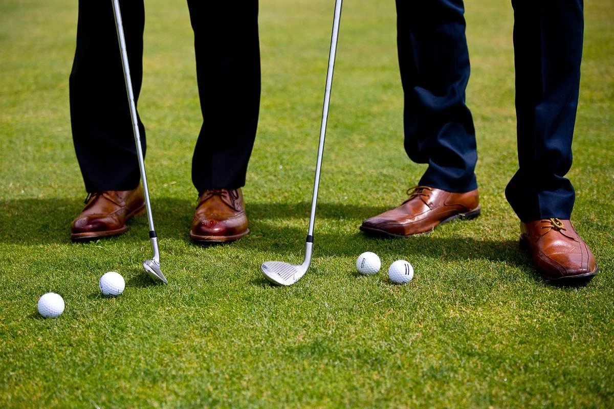 Two groomsmen in suits stand on a golf course, legs visible, holding clubs. Several golf balls are scattered on the grass, conveying a formal yet relaxed atmosphere during a Keystone Ranch wedding