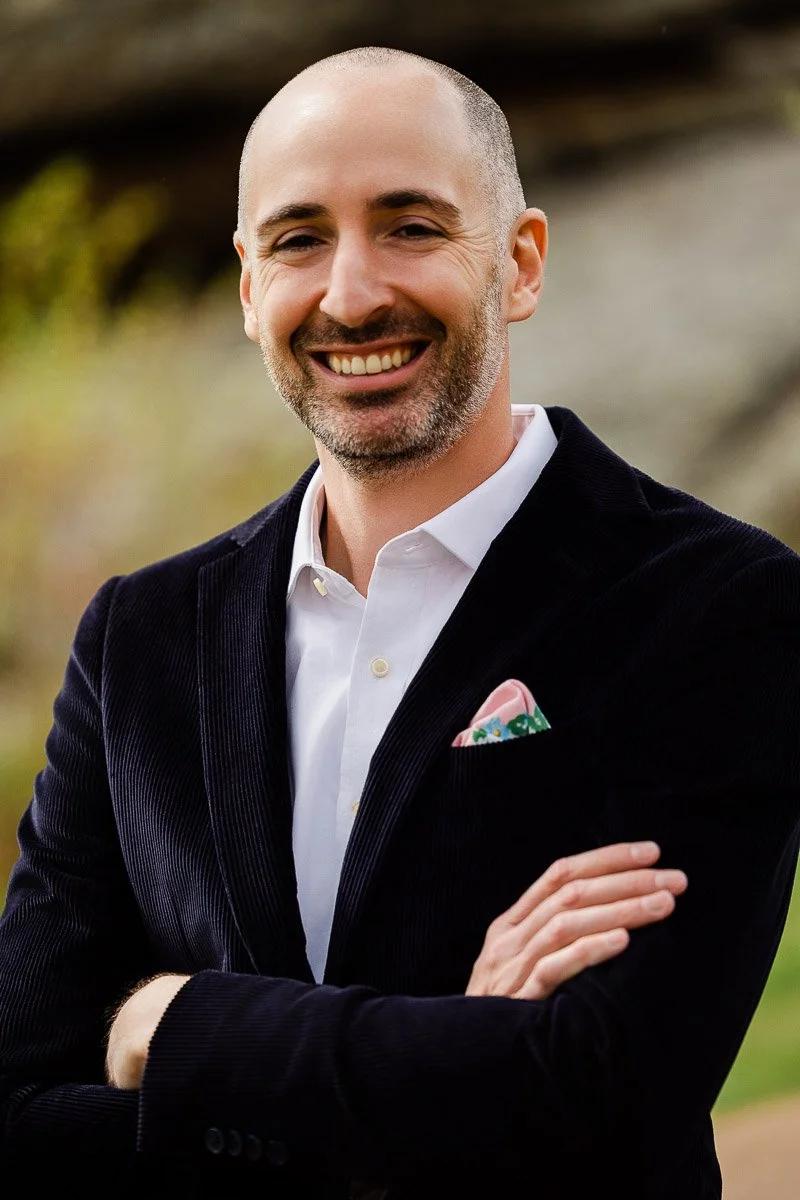Smiling man with a bald head wearing a dark blazer and white shirt, arms crossed. A colorful pocket square adds a stylish touch. Outdoor setting.