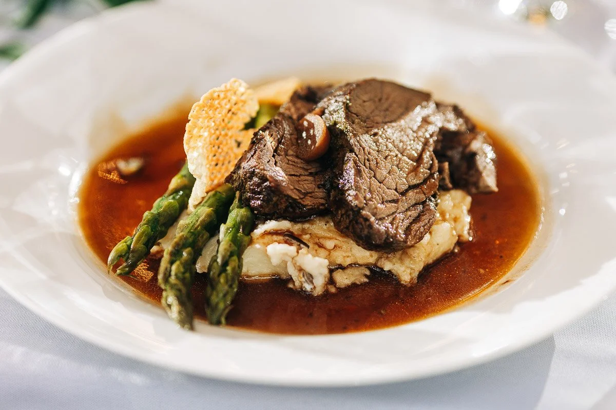 A gourmet dish featuring tender slices of beef on creamy mashed potatoes, with asparagus spears, and a delicate sauce in a white plate. Elegant presentation.