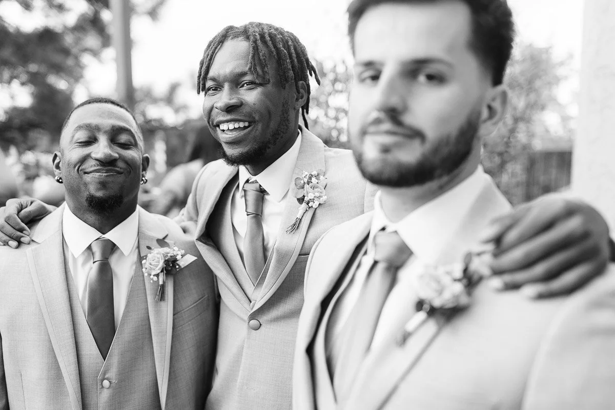Three groomsmen in light suits and ties smile warmly, embracing each other. The setting is outdoors, conveying joy and camaraderie at a wedding.