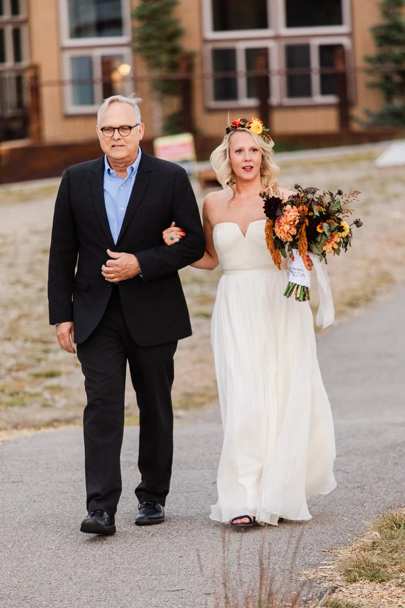 A bride in a flowing white dress and flower crown walks arm-in-arm with an older man in a dark suit. She holds a vibrant autumn bouquet, exuding joy during a Keystone ski mountain wedding.
