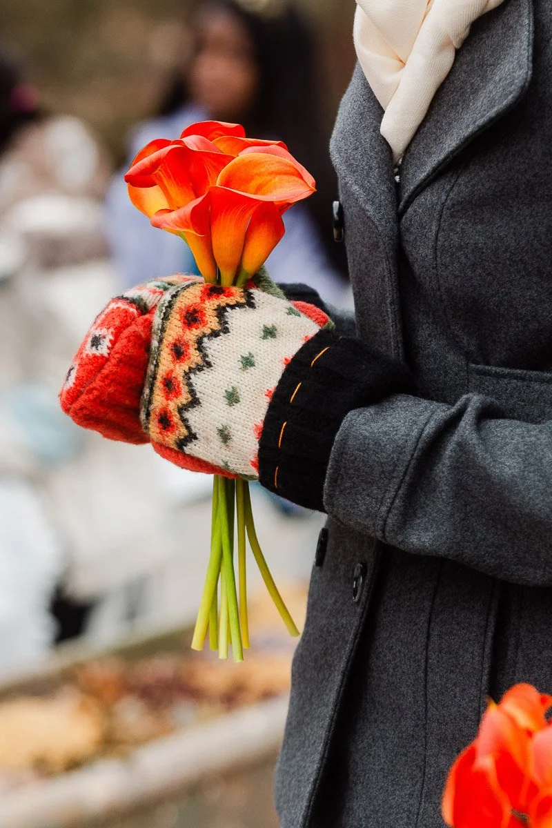 A person in a gray coat and patterned mittens holds vibrant orange calla lilies. The atmosphere feels warm and thoughtful, with soft background focus.