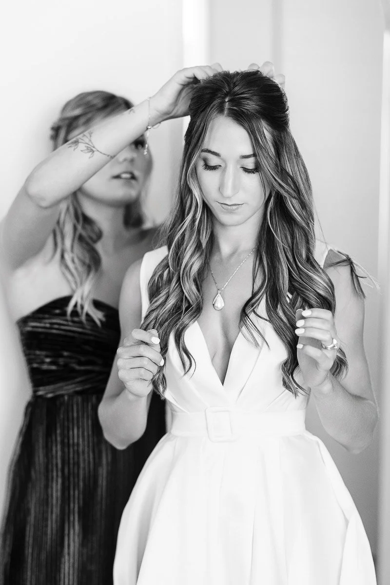 Bride in a white dress adjusts her hair while another woman in a dark strapless dress assists her. The scene conveys a calm, intimate moment.