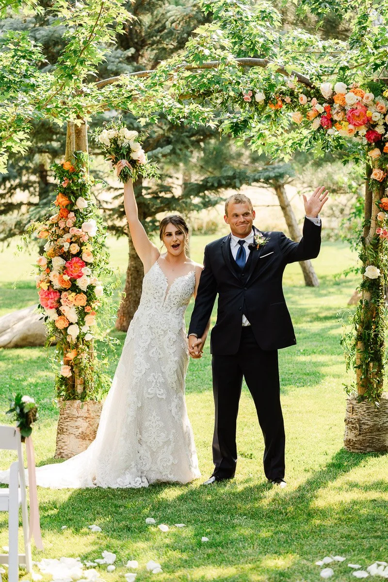 A joyful couple under a floral arch at their wedding, with the bride in a lace gown holding a bouquet, both waving joyously against a lush, sunny backdrop.