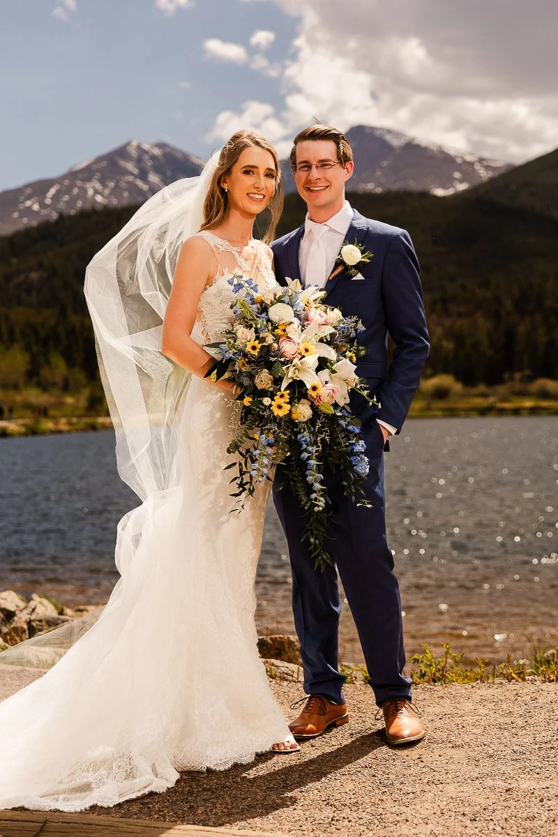 A smiling bride and groom stand by a tranquil Lily Lake with snow-capped mountains in the background. She wears a lace gown and holds a vibrant bouquet, while he is in a navy suit. The scene conveys joy and natural beauty.