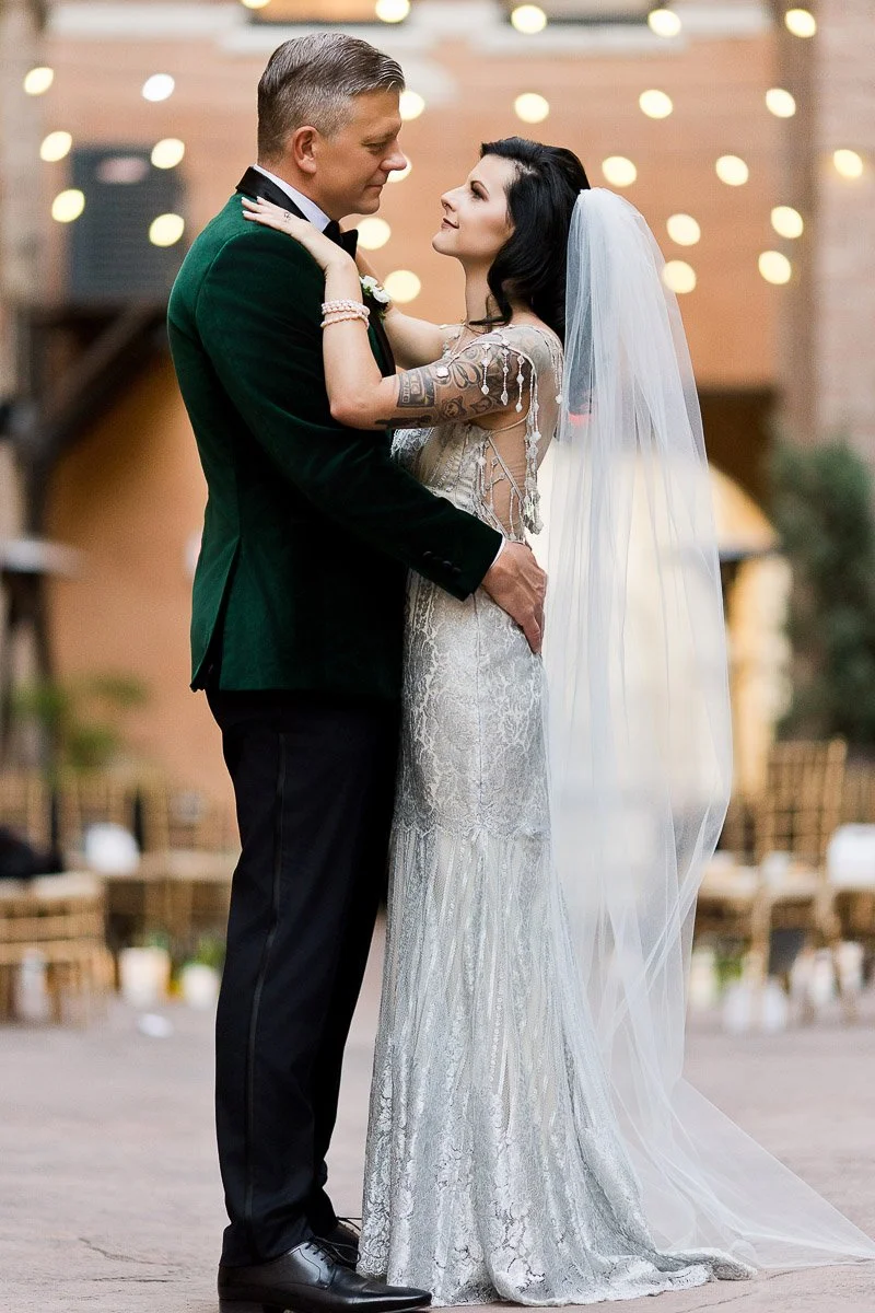 A couple embraces tenderly outdoors, the man in a dark suit and the woman in a white bridal gown with a veil. String lights twinkle softly above.