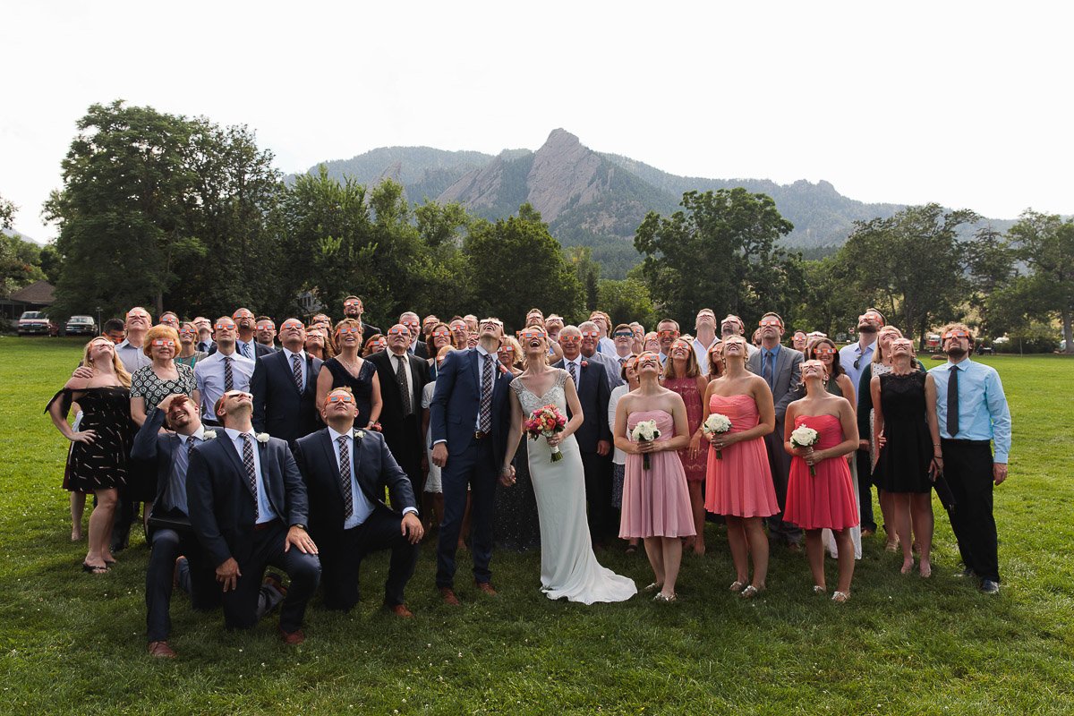 A large wedding group gathers outdoors on grass, all looking upward the solar eclipse eyewear with excitement. The bride in white holds flowers, bridesmaids in pink, against a mountain backdrop.