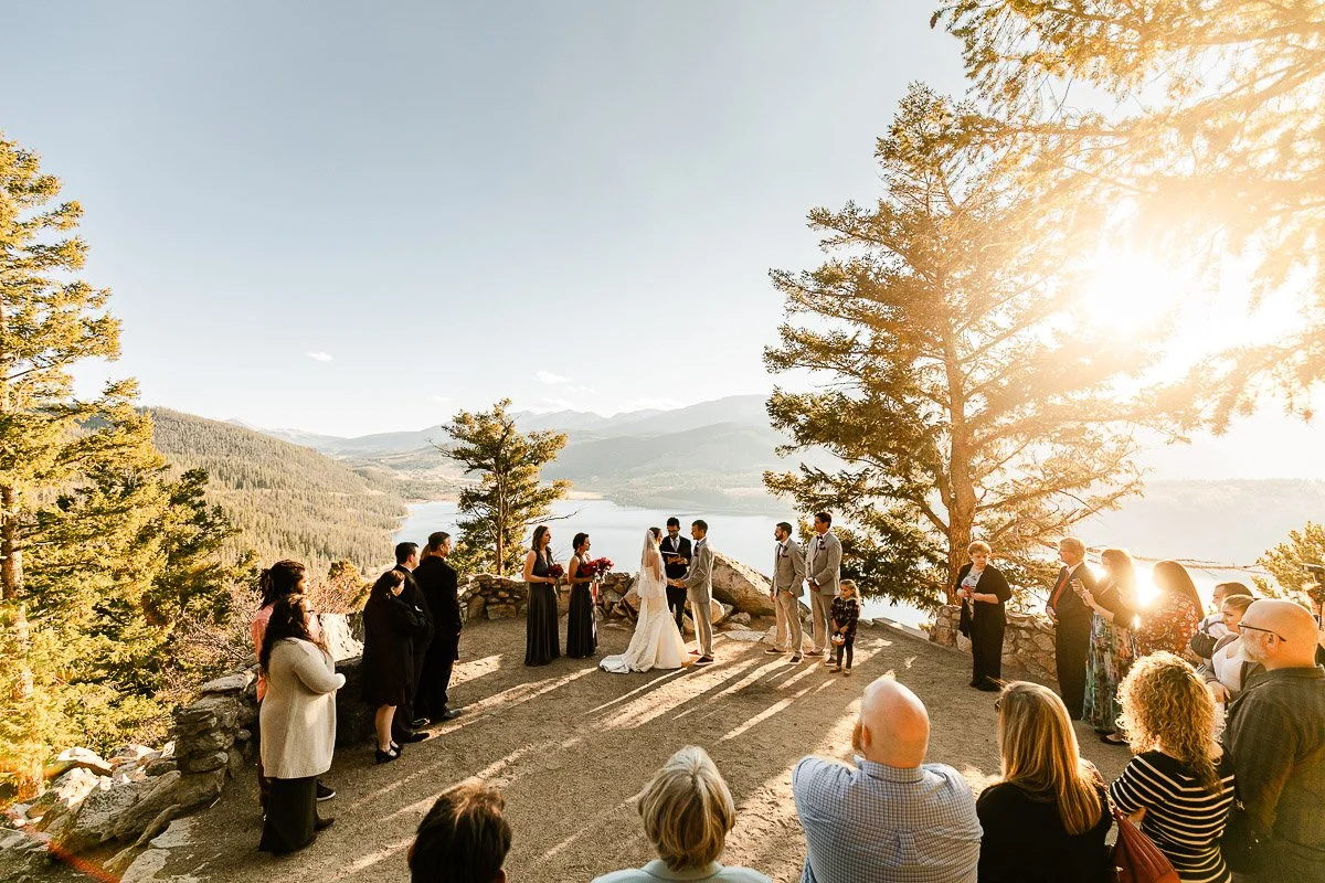 A wedding ceremony unfolds on a scenic mountainside with a view of a lake. Guests gather around the couple under a clear sky, with sunlight filtering through the trees, creating a warm, joyful ambiance.