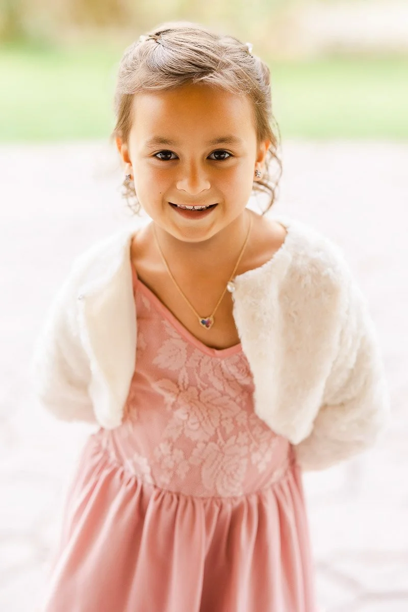 Young girl smiling, wearing a pink dress with floral lace and a white fur shrug. She has a heart necklace. Bright, cheerful outdoor setting.