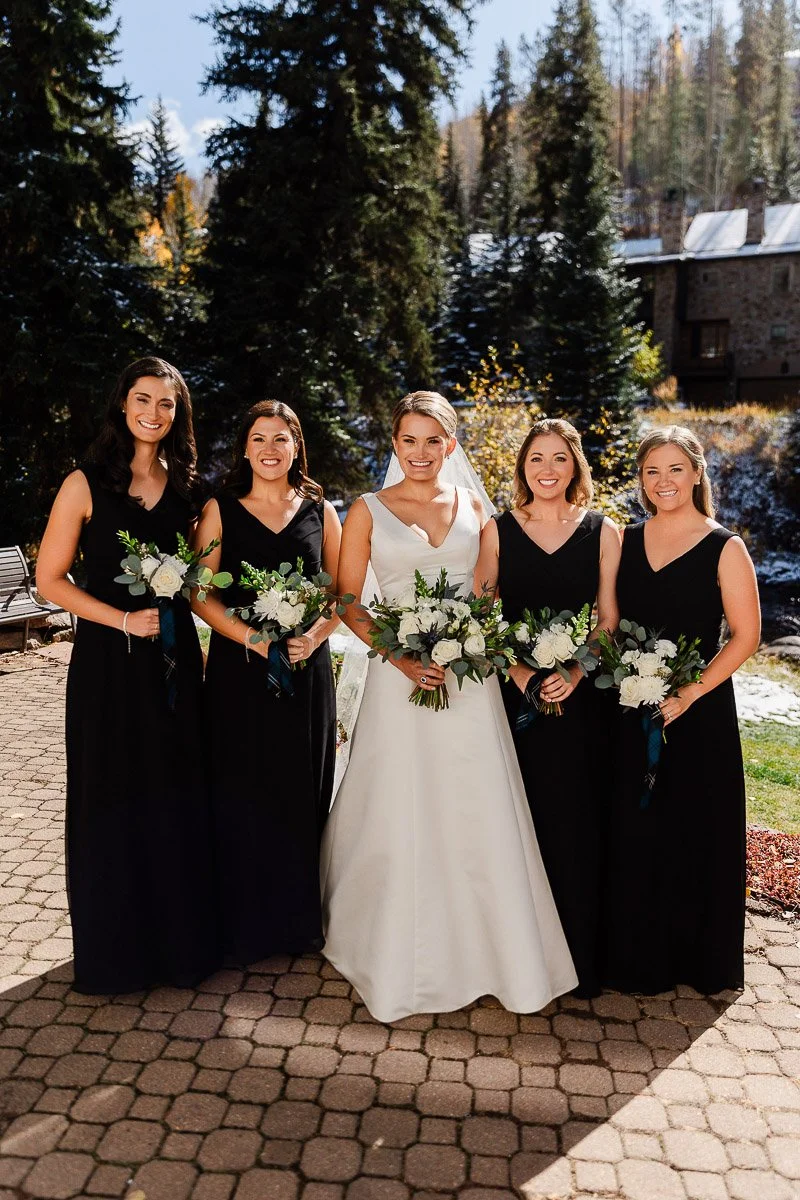 Bride in white gown smiles with four bridesmaids in black dresses, holding bouquets of white flowers. They stand on a sunlit path, surrounded by trees.