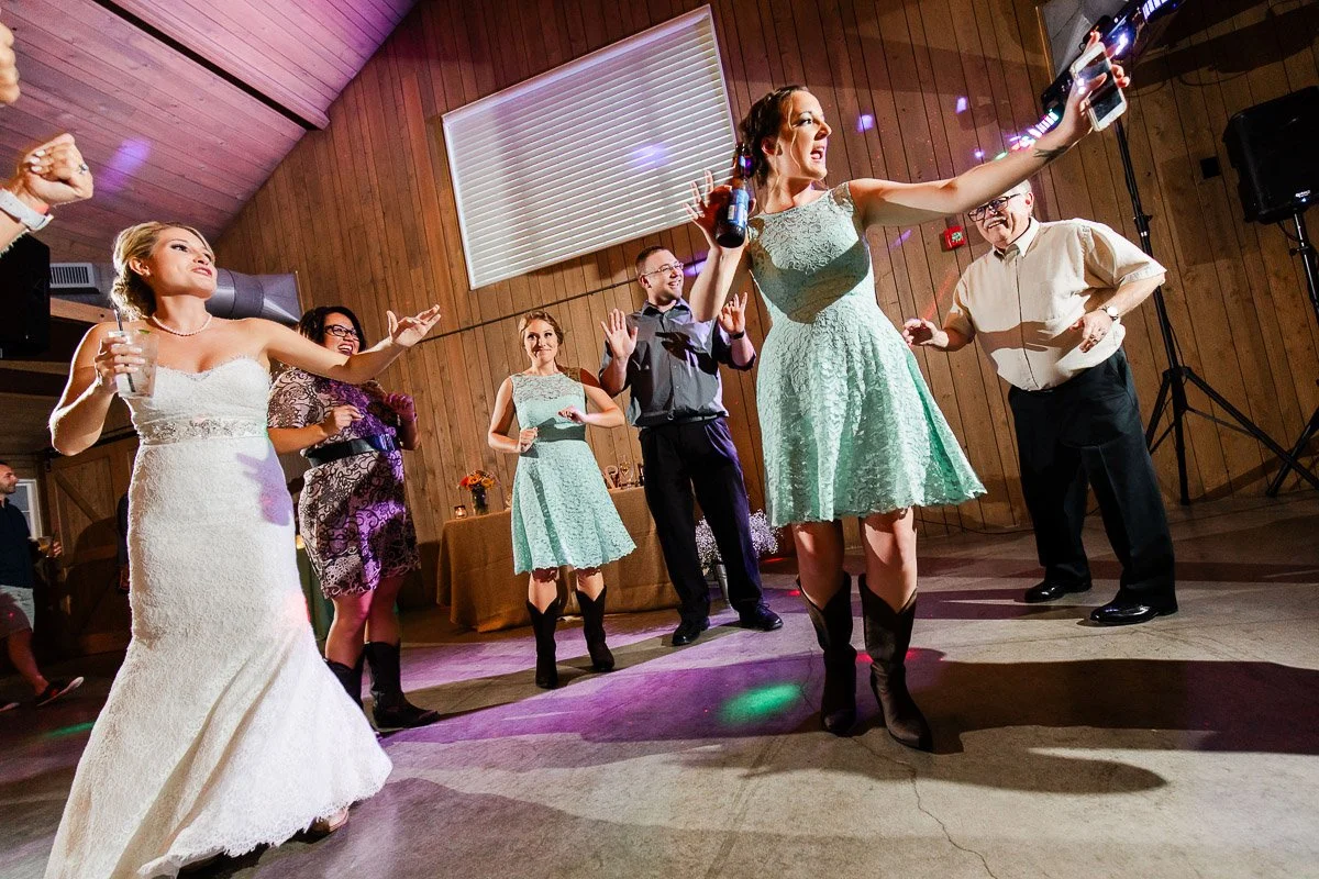 A bride and groom walk hand in hand across a grassy field at sunset, with a serene lake and cloudy sky in the background, creating a romantic atmosphere.