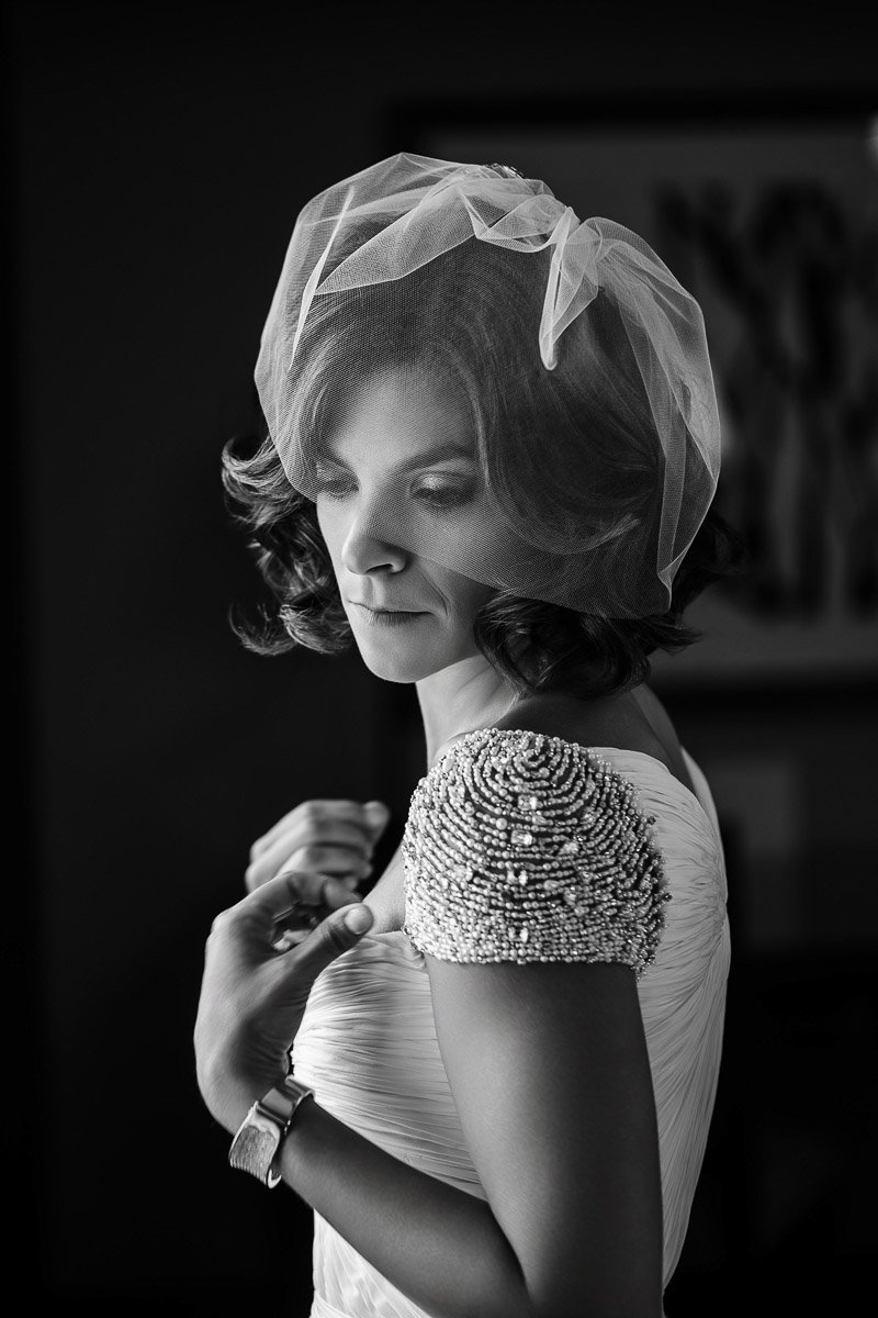 Elegant woman in a bridal gown with a veiled hat gazes downward, exuding a serene and contemplative mood. Her dress features intricate beaded details.