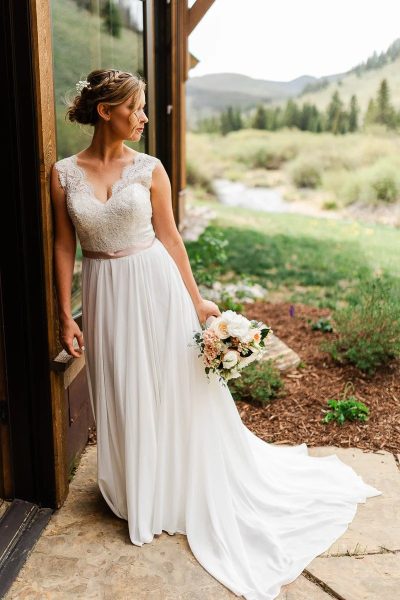 Bride in elegant lace wedding dress holds bouquet and leans against rustic building. Scenic backdrop features green hills and a stream, conveying tranquility.