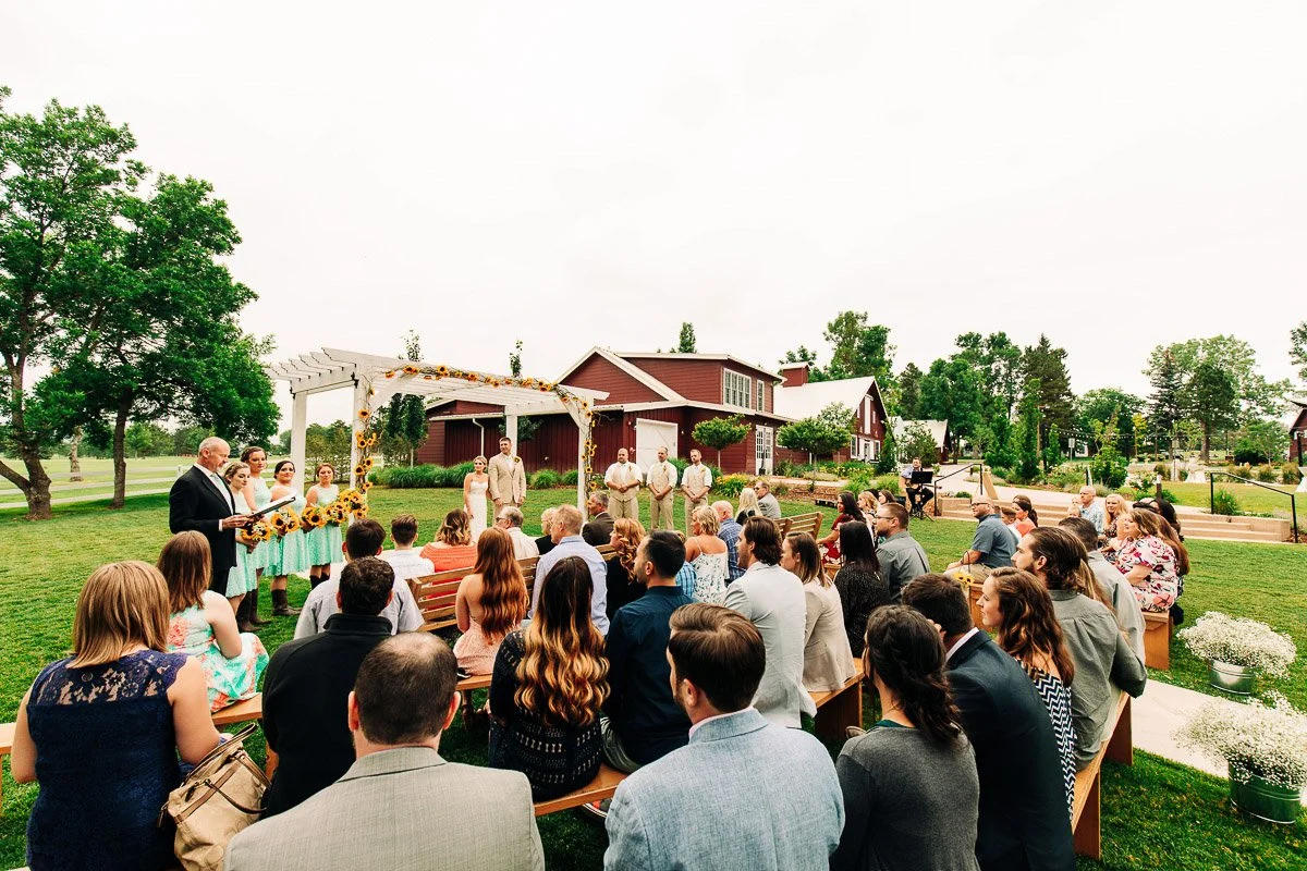 Outdoor wedding ceremony with guests seated on wooden benches, facing a flower-adorned arbor. Bridesmaids in aqua dresses and groomsmen in beige suits stand nearby. Rustic barn and lush greenery complete the serene setting.