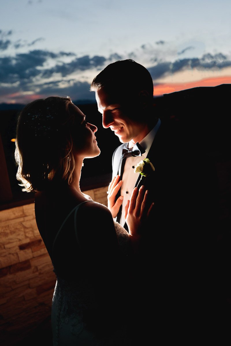 A silhouetted bride and groom gaze at each other tenderly, illuminated by soft sunset light. The scene conveys warmth and romance against a twilight sky.