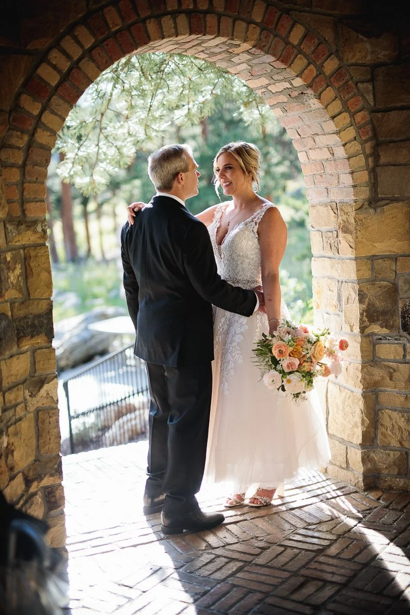 Bride and groom smile under a sunlit stone archway. She holds a bouquet of pink and white flowers, creating a romantic and joyous atmosphere.