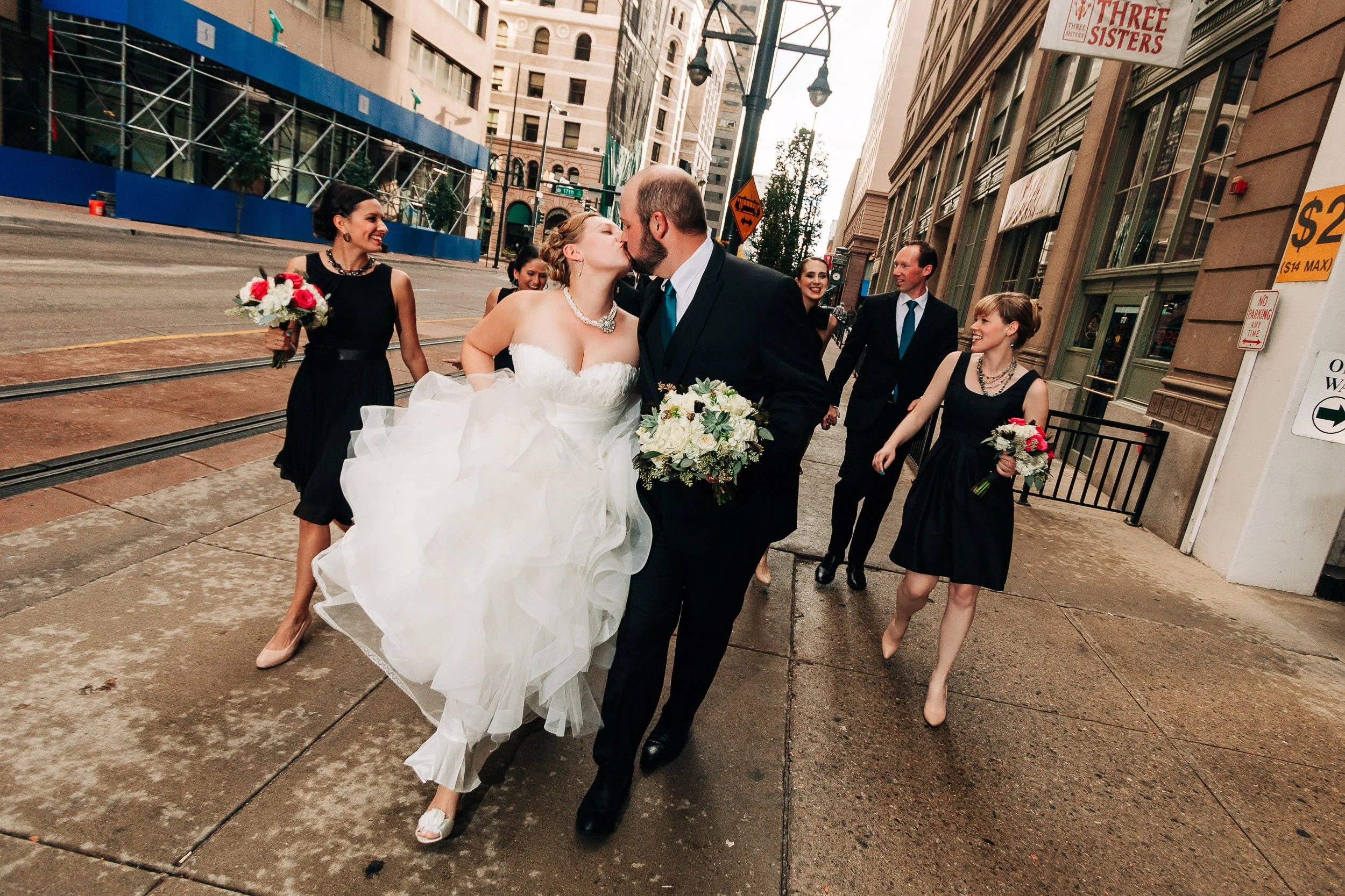 Bride and groom kiss on city streets followed by bridesmaids and groomsmen during a Magnolia Hotel wedding in Denver, Colorado