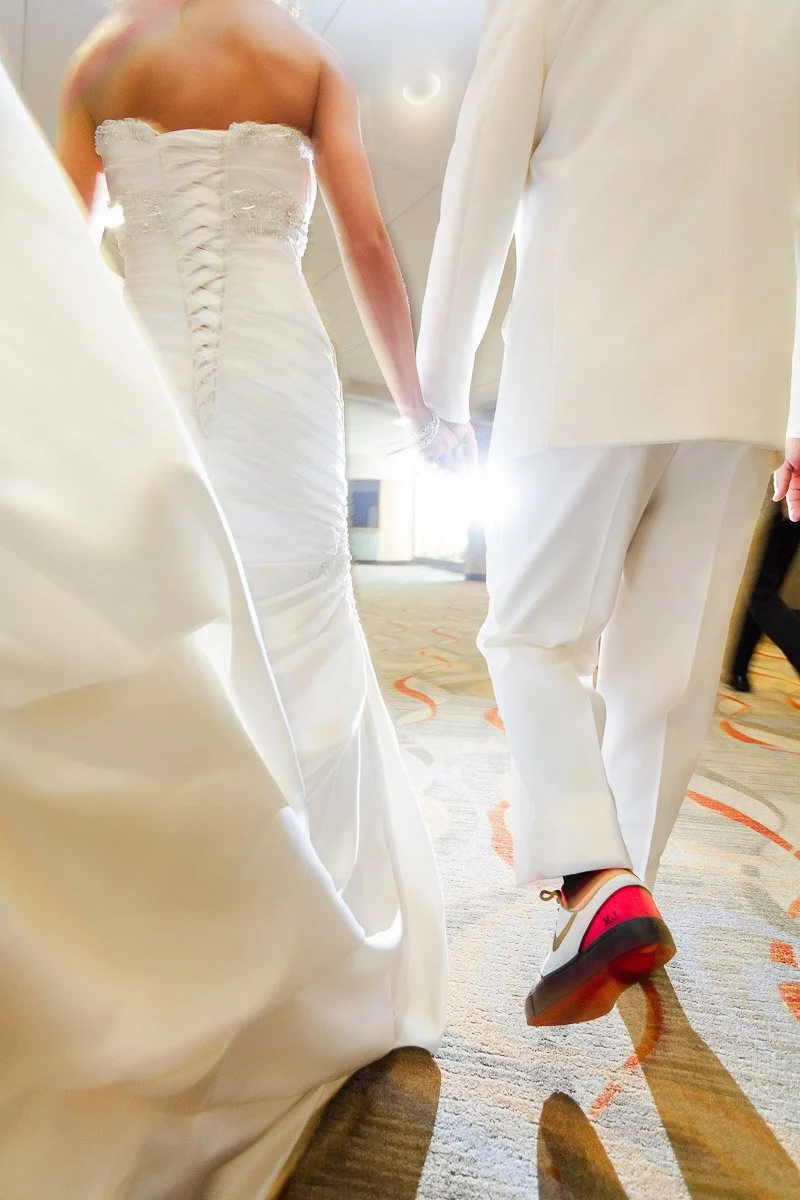 A couple dressed in elegant wedding attire, seen from behind, walk hand in hand. The bride wears a white gown and the groom wears a white suit with pink-soled shoes. The atmosphere is bright and joyful.
