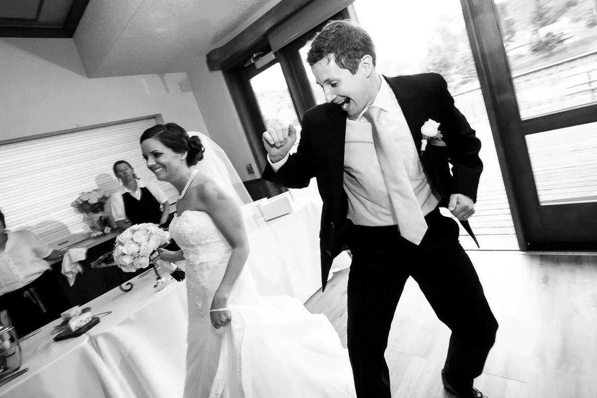 A joyful bride and groom dance indoors. The bride, in a white gown, holds a bouquet, smiling. The groom, in a suit, appears to be celebrating energetically.