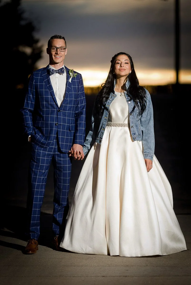A couple stands hand in hand at sunset. The man wears a blue checkered suit, and the woman wears a flowing white gown with a denim jacket. They appear joyful and content.