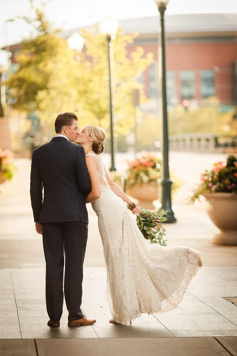 A newlywed couple shares a kiss in a sunlit, urban setting. The bride in a lace dress holds a bouquet, while the groom wears a dark suit. Romantic and joyful during a Coohills Restaurant wedding in Denver, Colorado.