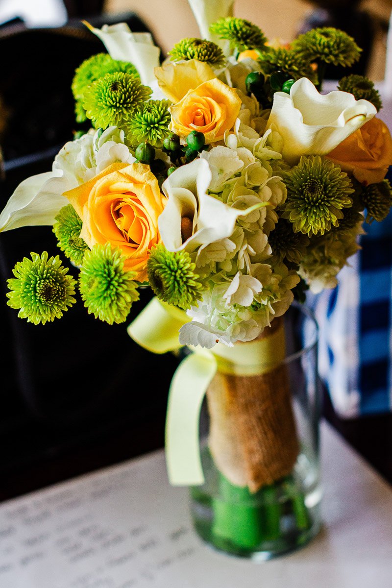 A vibrant bouquet featuring yellow roses, white lilies, and green chrysanthemums in a glass vase, with a rustic burlap wrap, on a dark surface.