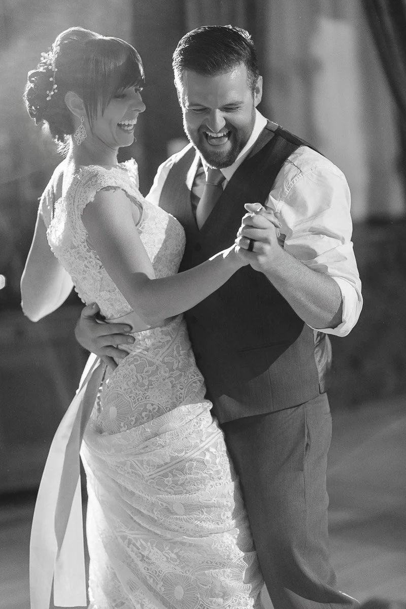 A joyful couple dances, the bride in a lace gown and the groom in a vest. Both are smiling and laughing, exuding happiness in a softly lit setting.