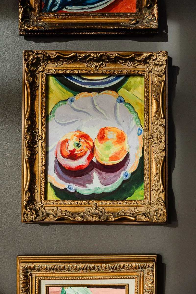 Framed painting of two apples on a white plate with a vibrant green background. The ornate gold frame adds a classic touch, capturing a warm, artistic ambiance.