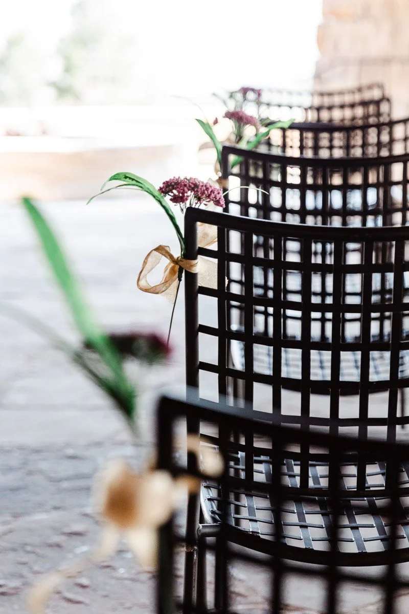 A row of black metal chairs decorated with purple flowers and green leaves tied with beige ribbon, set on a stone pathway in soft natural light.