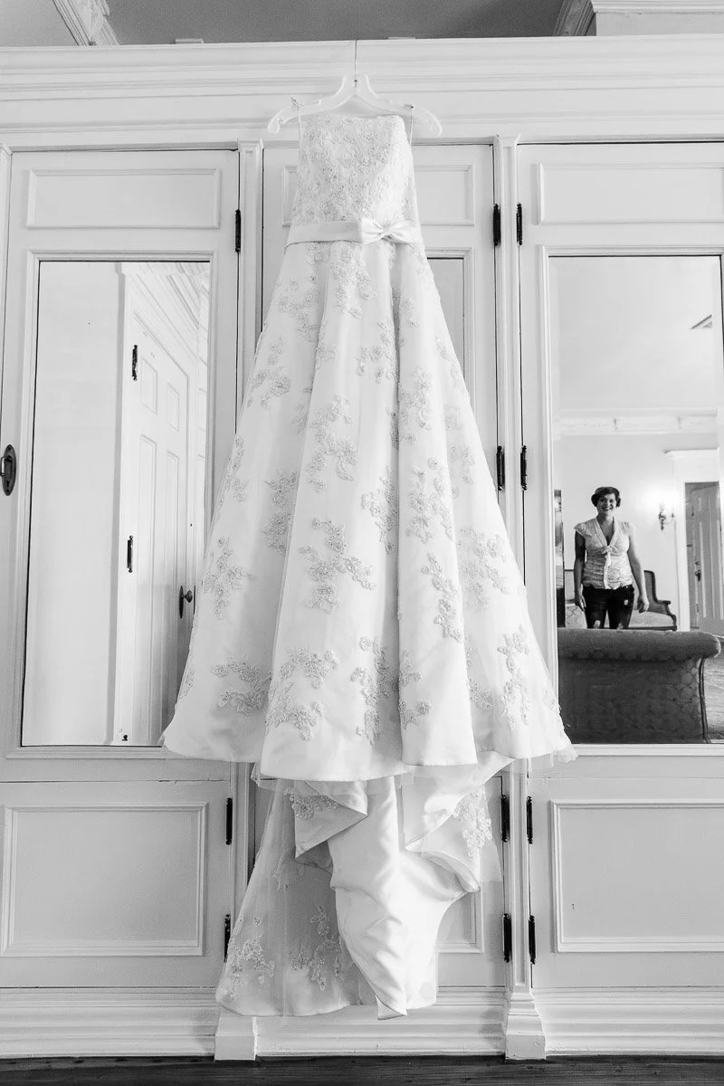 A wedding dress with intricate lace patterns hangs elegantly on a wardrobe. In the mirror reflection, a person smiles warmly, adding a joyful tone.