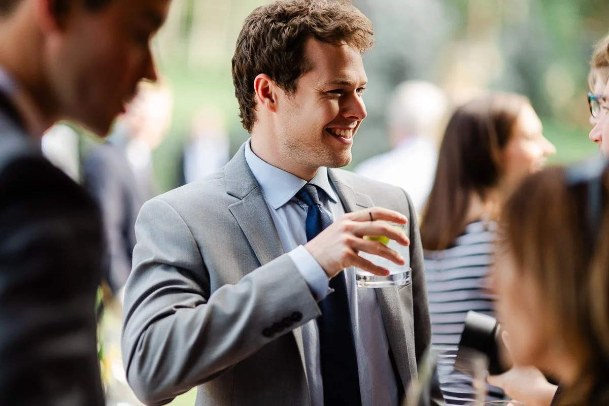 A man in a gray suit, holding a drink, smiles during an outdoor event. Blurred people in background suggest a lively, social atmosphere.