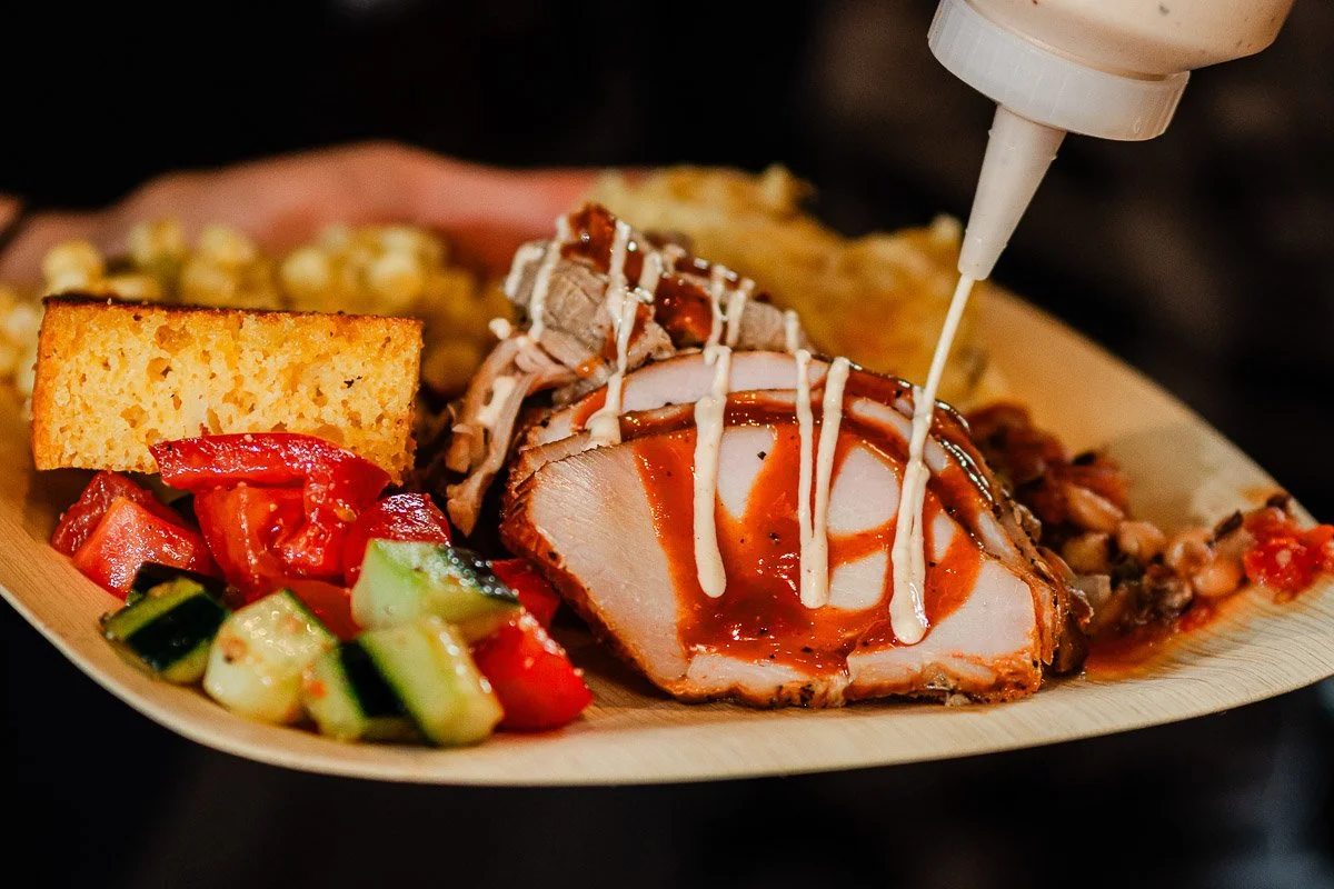 Plate with sliced grilled meat drizzled with sauce, cornbread, cucumber-tomato salad, beans, and mac and cheese. Appealing and vibrant.