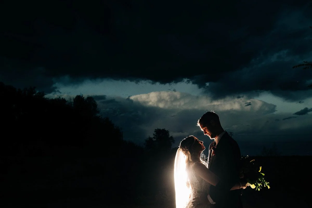 Silhouetted bride and groom embrace under a dramatic, cloudy night sky. A soft light illuminates them, creating a romantic and intimate mood.