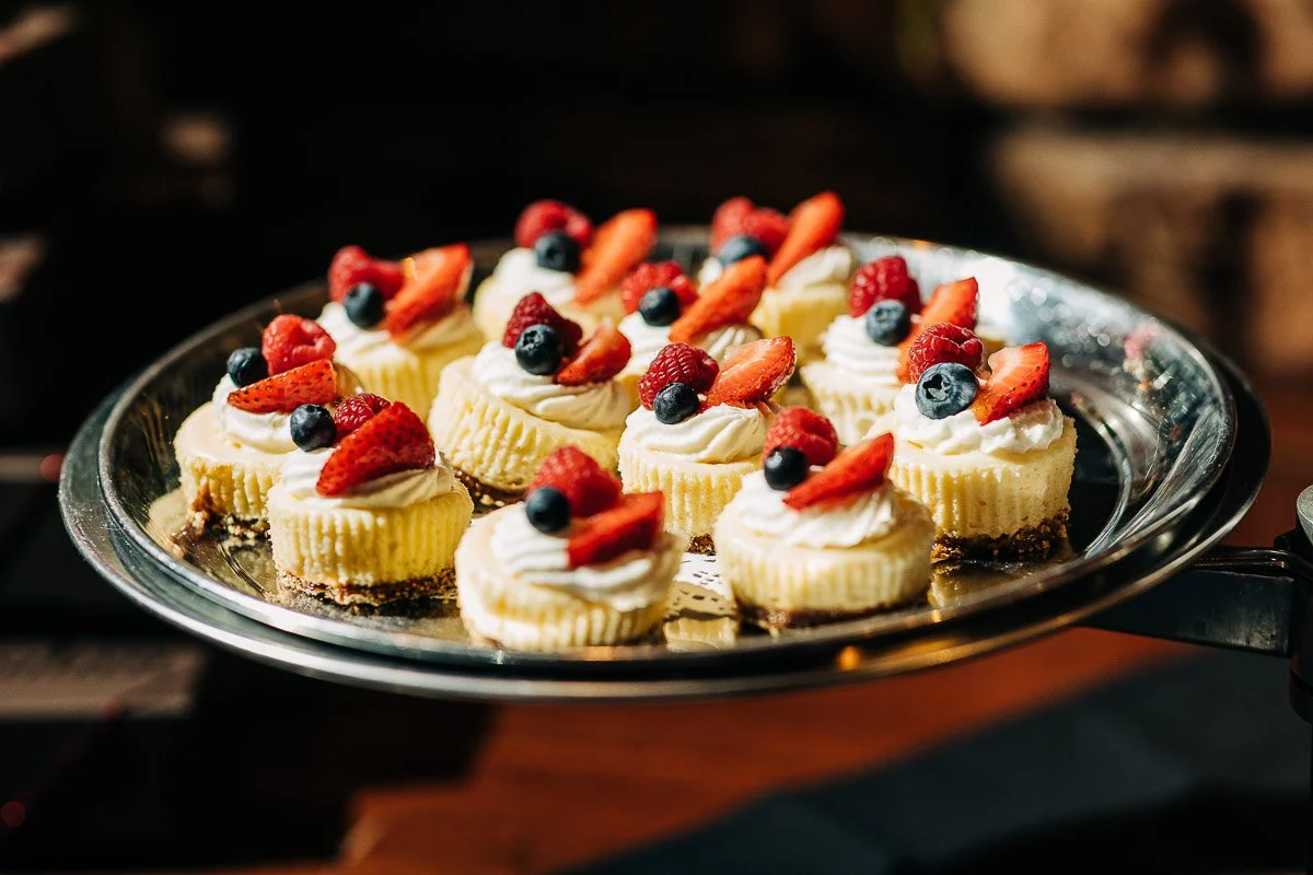 Mini cheesecakes topped with fresh strawberries, raspberries, and blueberries are artfully arranged on a silver tray, bathed in warm sunlight.