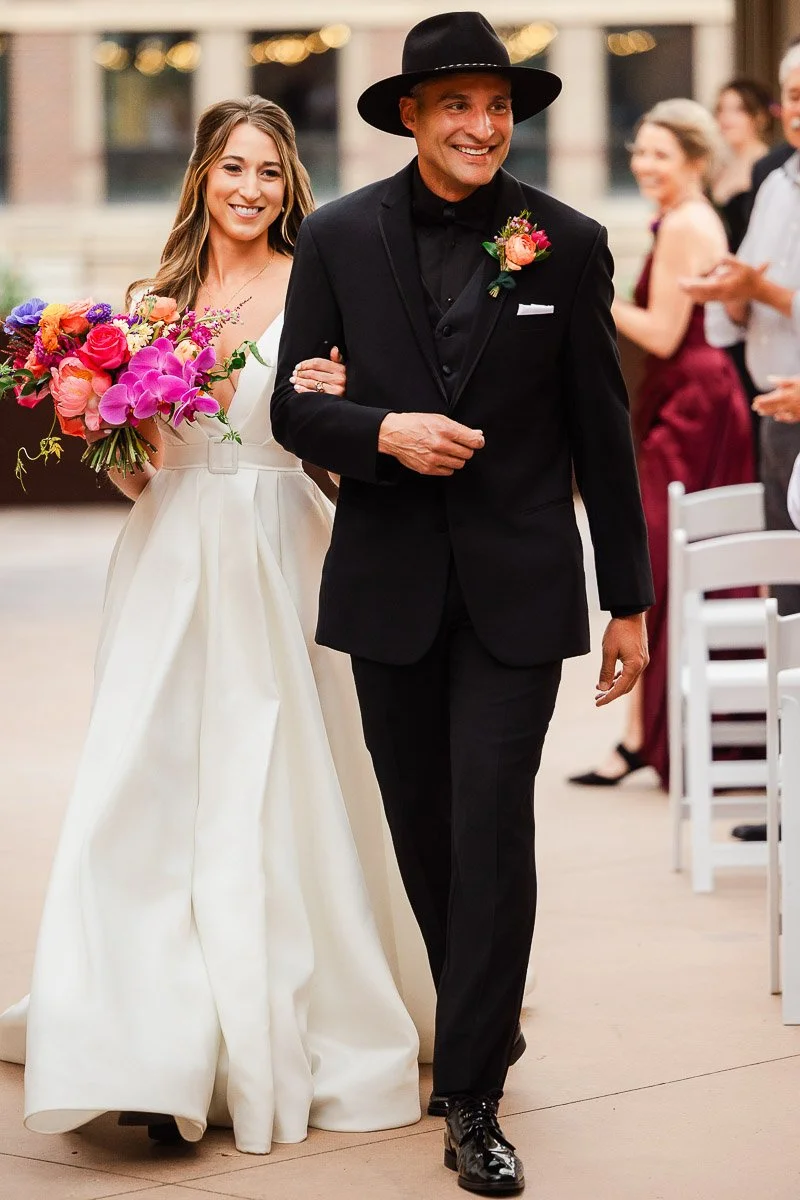 Bride in a white gown and man in a black suit with a hat walk arm in arm down an aisle. She holds a vibrant bouquet, both smiling joyfully.