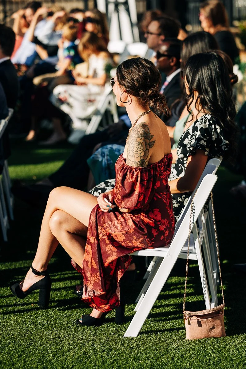 A woman in a red floral dress and black heels, showcasing a tattoo, sits on a white folding chair outdoors. The setting is sunny, with an audience in the background.